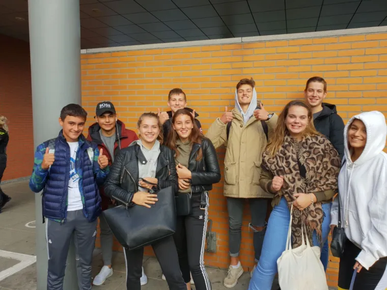 Groep cursisten van de theoriedagcursus binnen voor een gele muur, met duimen omhoog.
