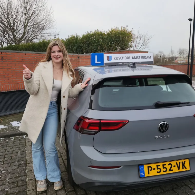Geslaagde leerling met duim omhoog naast de grijze Golf met het dakbord van Rijschool Amsterdam.