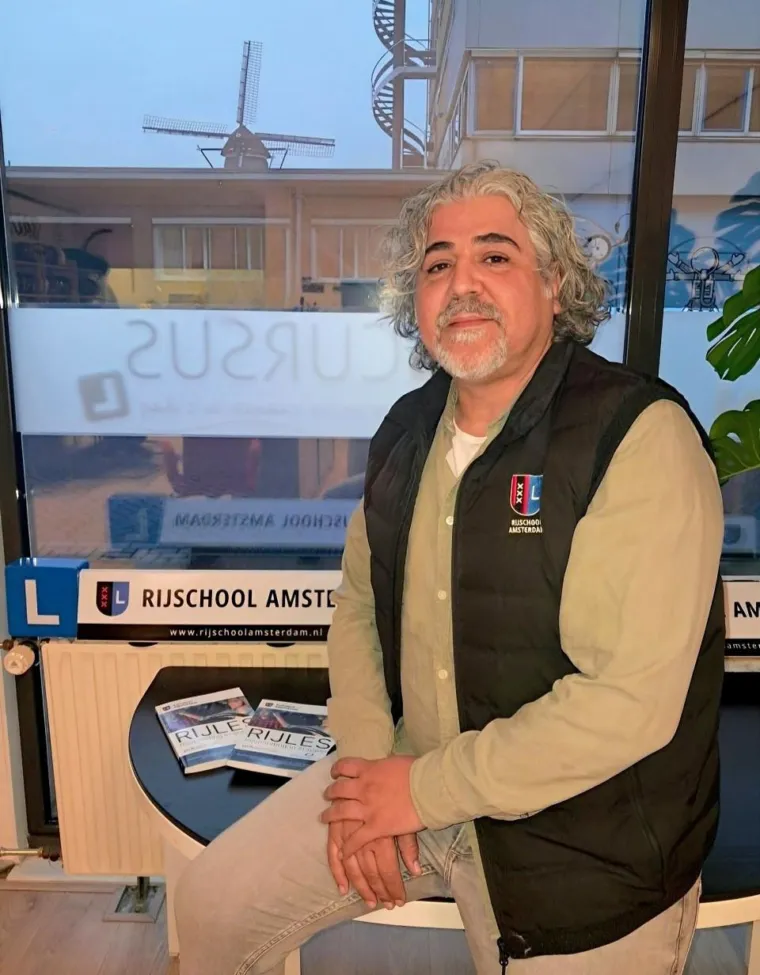 Rijinstructeur Hassan Laboui zit in het lokaal van Rijschool Amsterdam, met het dakbord en een windmolen achter het raam.